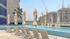 A poolside area with lounge chairs and umbrellas, a view of a construction site, and a modern building in the background. - Silkhaus Luxe 3BR Apartment by the Canal (Dubai)