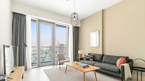 A modern living room with a sofa, a coffee table, a TV, and a view of a construction site through large windows. - Silkhaus Contemporary 2BR Canal View Stay (Dubai)