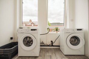 A laundry room with two front-loading washing machines, a black laundry basket, and a window with a view of buildings outside. - 1 Bedroom Studio Apartment ~ Central Middlesbrough (Middlesbrough)