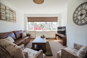 A living room with a brown leather sofa, a wooden coffee table, a television on a wooden stand, and a large wall clock. - Stylish 3BR Bungalow with Garden | Sleeps 6 (Redcar and Cleveland)