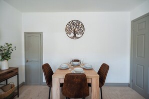 A dining area with a wooden table set for four, two chairs, a decorative wall piece, and a door. - Stylish 3BR Bungalow with Garden | Sleeps 6 (Redcar and Cleveland)