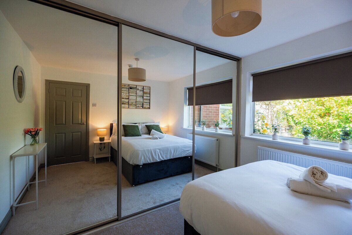 A bedroom with a large mirror, a bed with white linens, a bedside table with a lamp, and a window with a view of greenery.