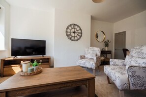 A living room with a flat-screen TV, a wooden coffee table, two patterned armchairs, a round wall clock, and a mirror. - Stylish 3BR Bungalow with Garden | Sleeps 6 (Redcar and Cleveland)
