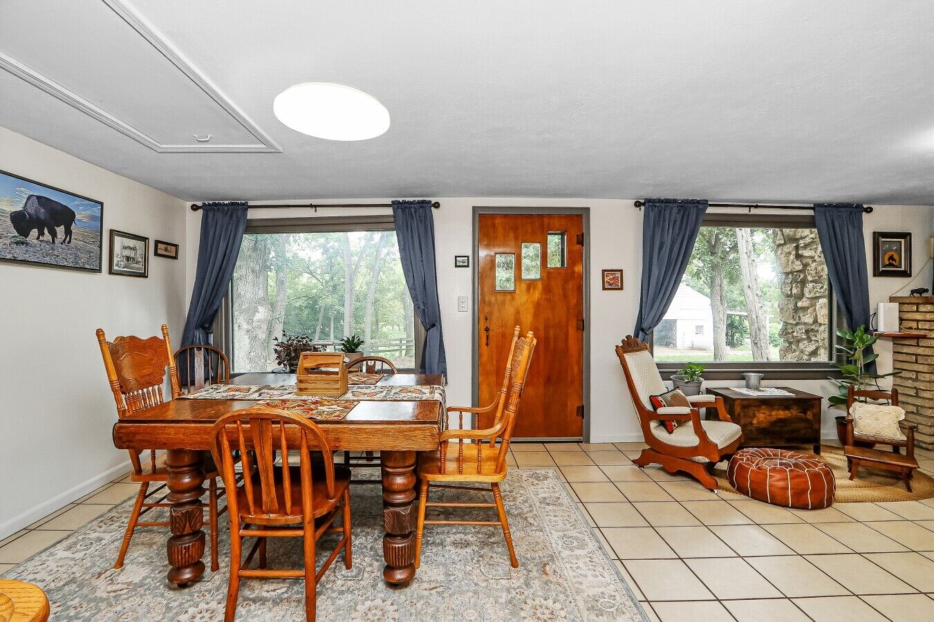 A dining area with a wooden table and chairs, a rug, and a view of trees through large windows.