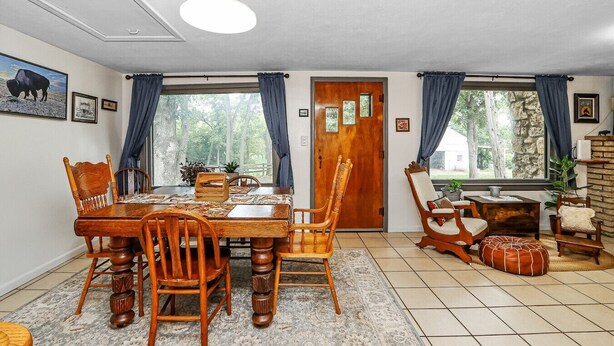 A dining area with a wooden table and chairs, a rug, and a view of trees through large windows.
