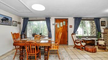 A dining area with a wooden table and chairs, a rug, and a view of trees through large windows.