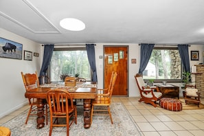 A dining area with a wooden table and chairs, a rug, and a view of trees through large windows. - Peaceful Lake Cabin 3BD - Surrounded by Nature (Manhattan)