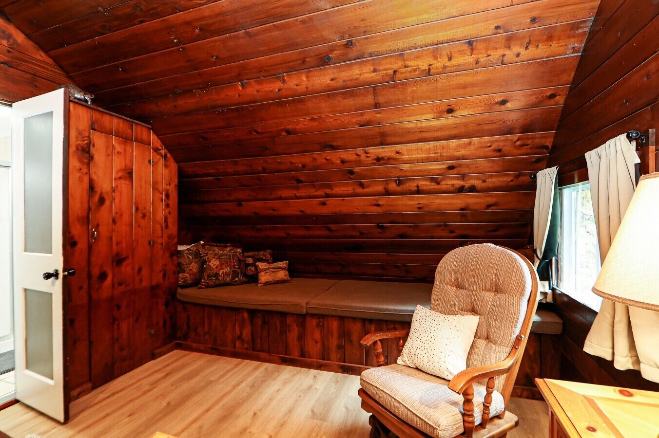 A cabin interior with a wooden ceiling, a bench, a cushioned chair, a table, and a door.