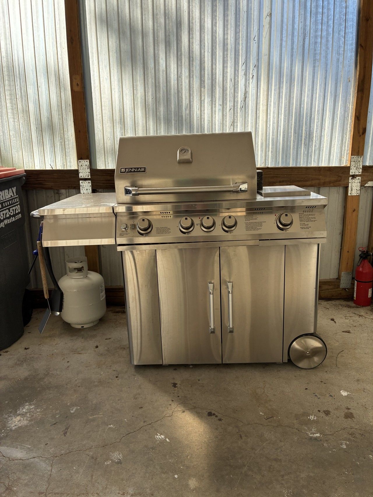 A stainless steel Jenn-Air gas range with four burners and a griddle, located in a garage with a corrugated metal wall and a concrete floor.