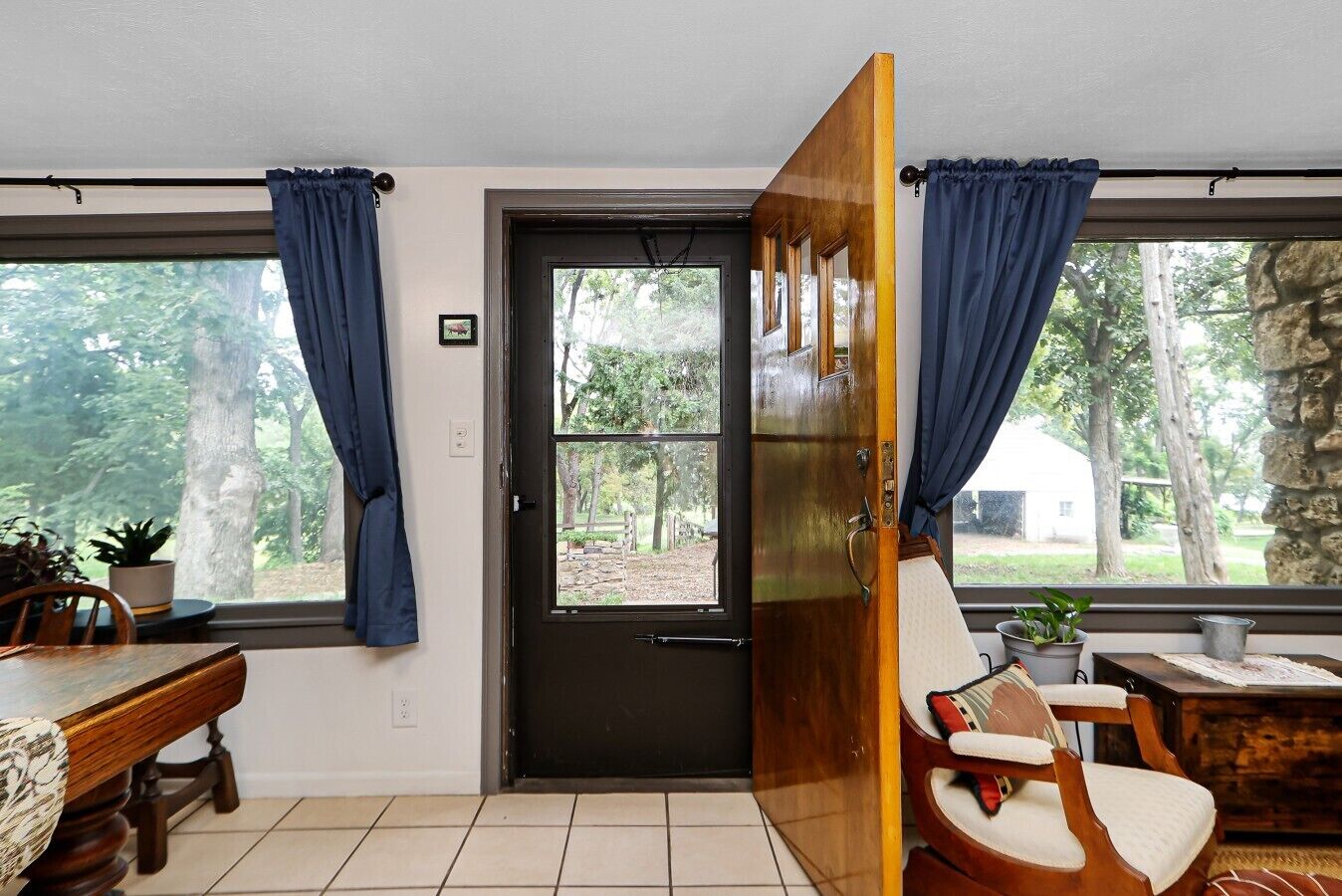 A room with a wooden door, a dining table, chairs, and a view of trees through the windows.