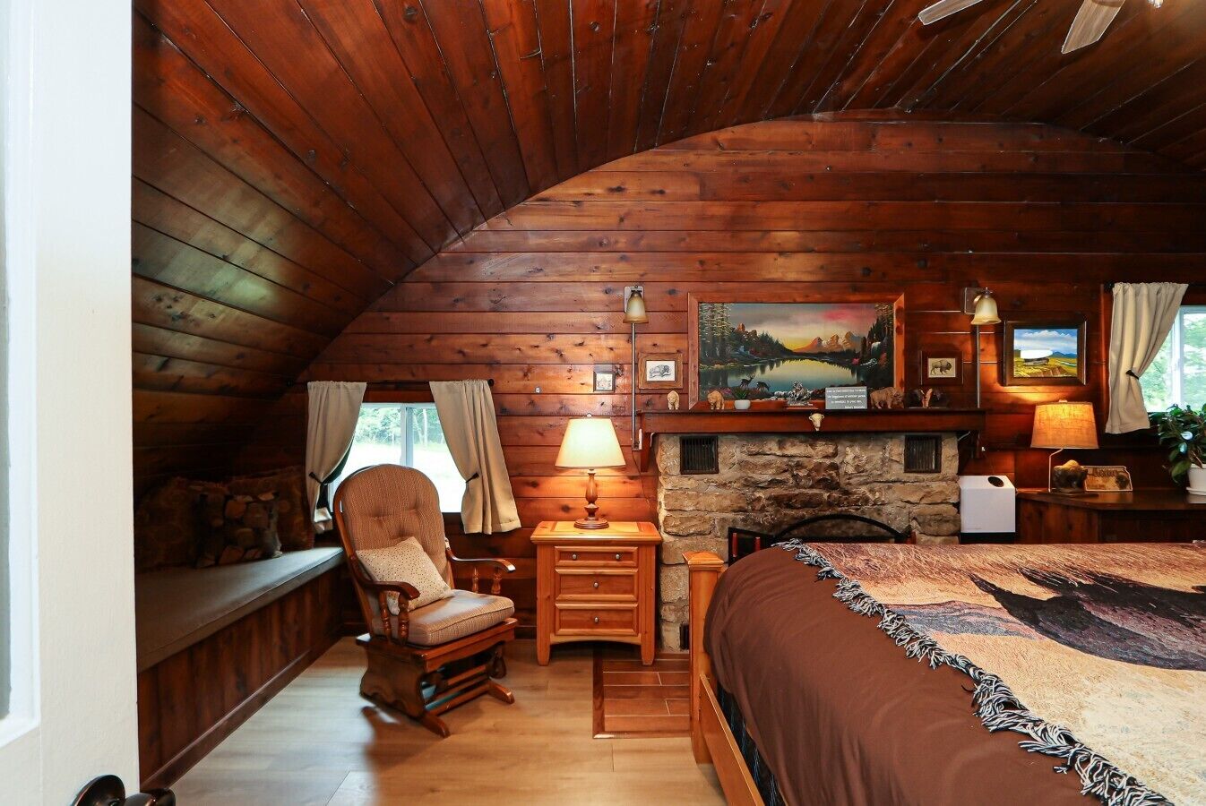 Une chambre confortable, aménagée dans le style d’un chalet, avec un plafond en bois, une cheminée en pierre, un lit recouvert d’une courtepointe à motifs, un fauteuil à bascule et un bureau avec une lampe.