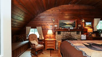 Une chambre confortable, aménagée dans le style d’un chalet, avec un plafond en bois, une cheminée en pierre, un lit recouvert d’une courtepointe à motifs, un fauteuil à bascule et un bureau avec une lampe.