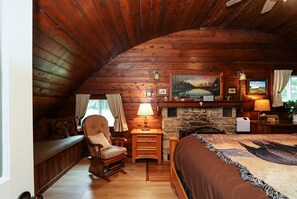 A cozy cabin-style bedroom with a wooden ceiling, a stone fireplace, a bed with a patterned bedspread, a rocking chair, and a desk with a lamp.