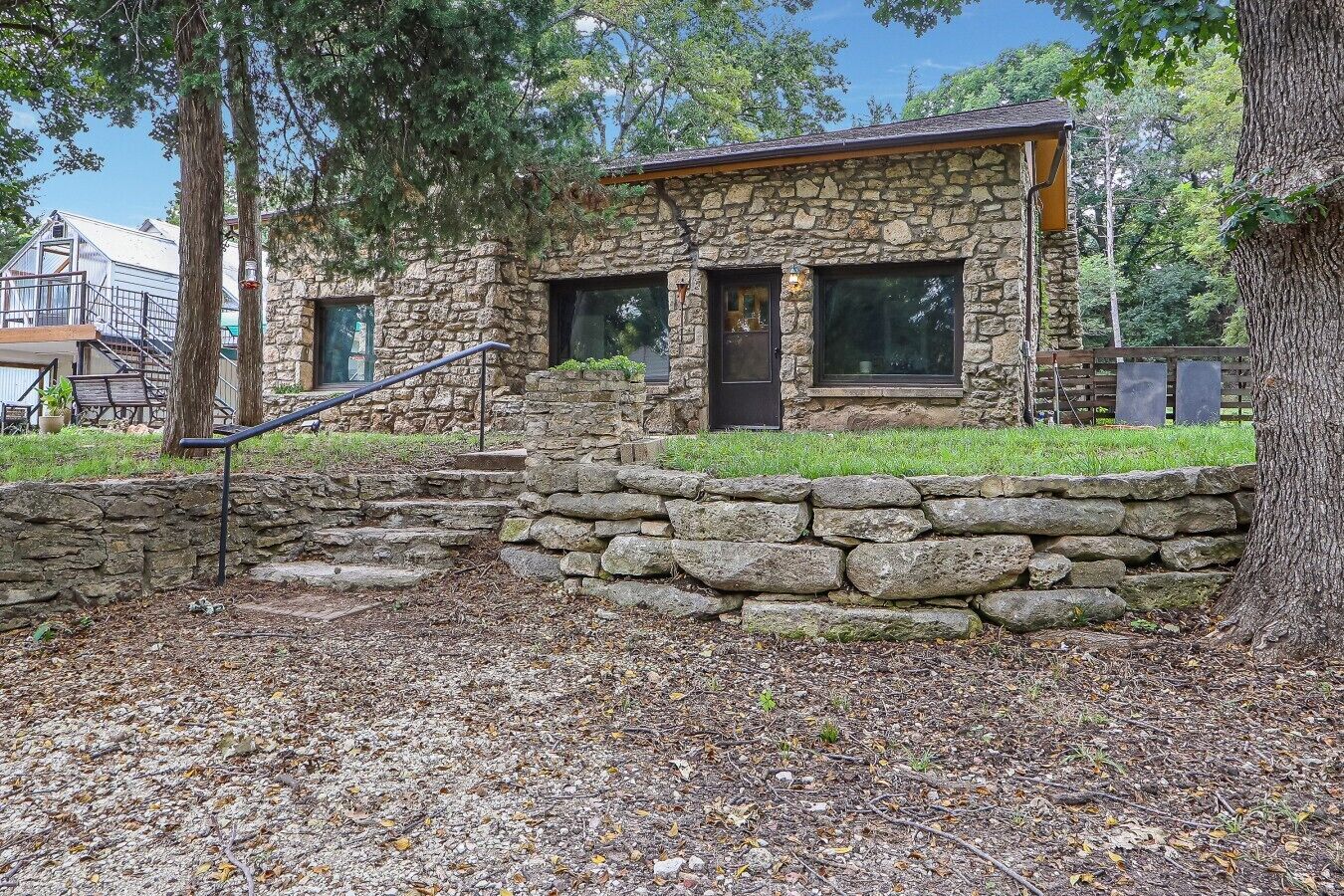 A stone house with a wooden staircase leading to a front door, surrounded by trees and a stone wall.