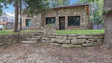 A stone house with a wooden staircase leading to a front door, surrounded by trees and a stone wall.