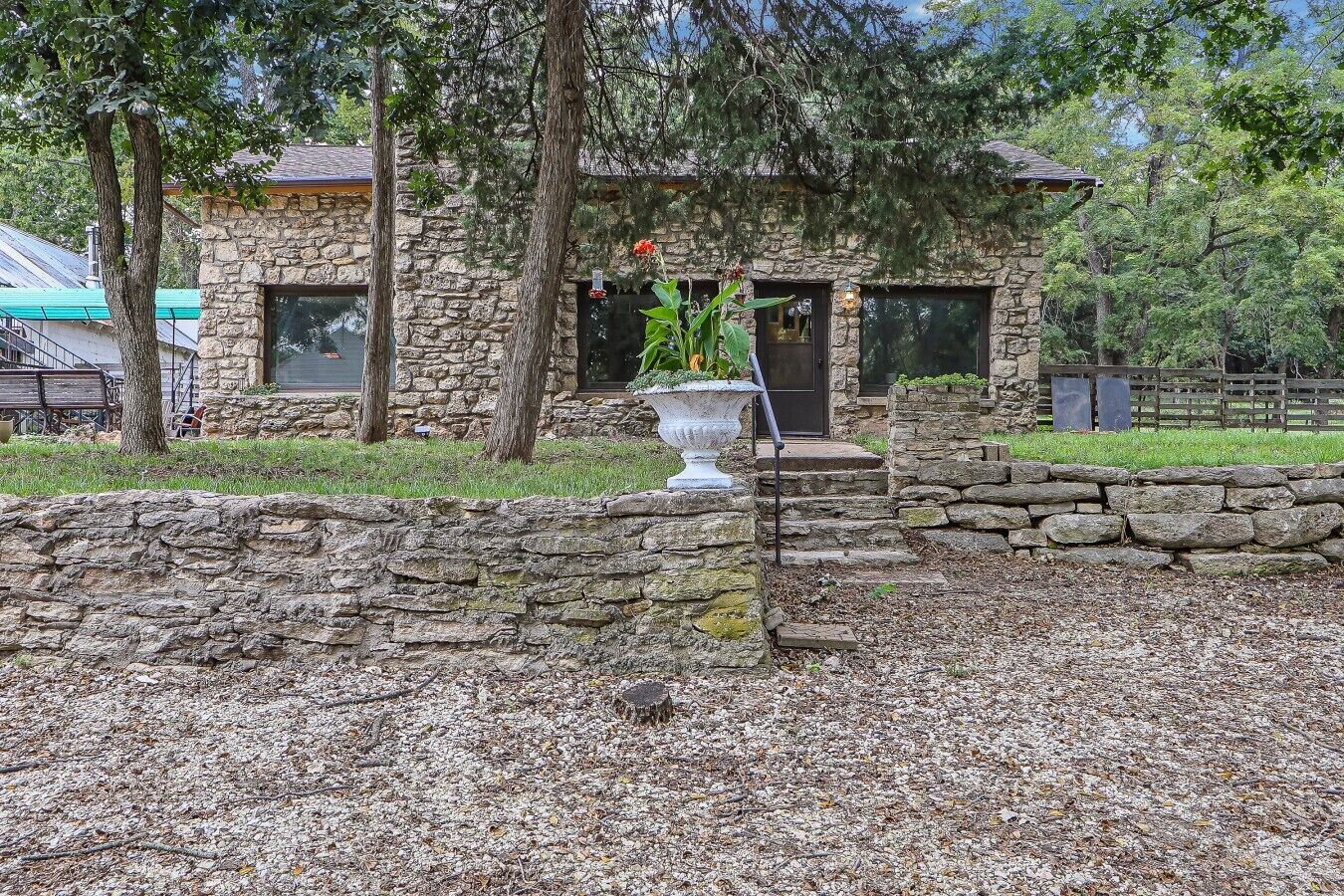 A stone building with a stone wall in front, a large planter with plants, and a wooden fence in the background.