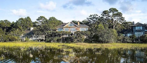 A large house with a reflection in a body of water, surrounded by trees and a grassy area.