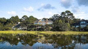 Uma casa grande refletida na água, rodeada por árvores e um gramado.
