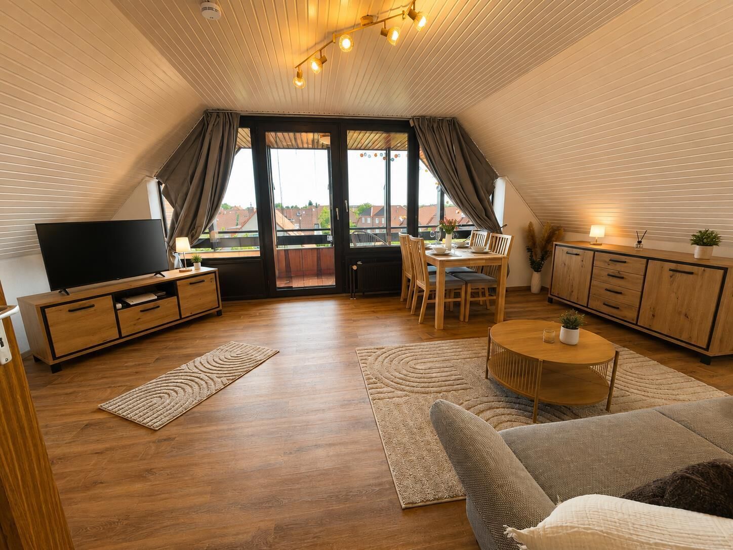 En modern lägenhet i loftstil med en platt-TV, möbler i trä, en matplats och stora fönster med utsikt över staden.