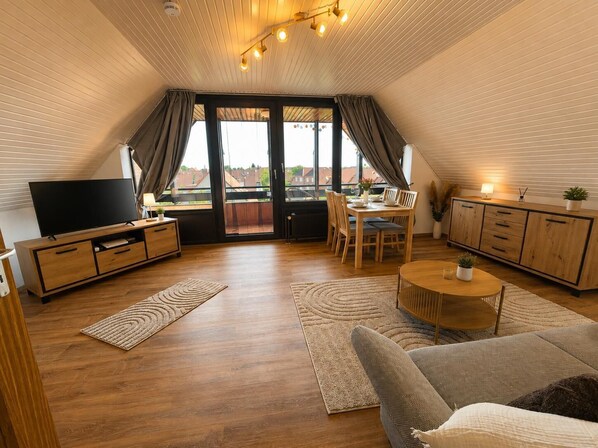 A modern loft apartment with a flat-screen TV, wooden furniture, a dining area, and large windows offering a city view. - Wolkendeck Vacation Rental (Hooksiel)
