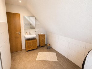 A bathroom with a wooden door, a white sink, a mirror, a cabinet, and a small trash bin. - Wolkendeck Vacation Rental (Hooksiel)