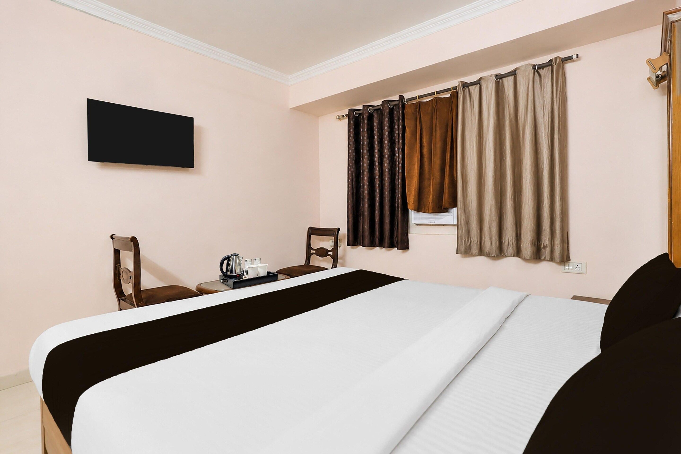A hotel room with a bed, a TV mounted on the wall, a window with curtains, and a small table with a kettle and cups.