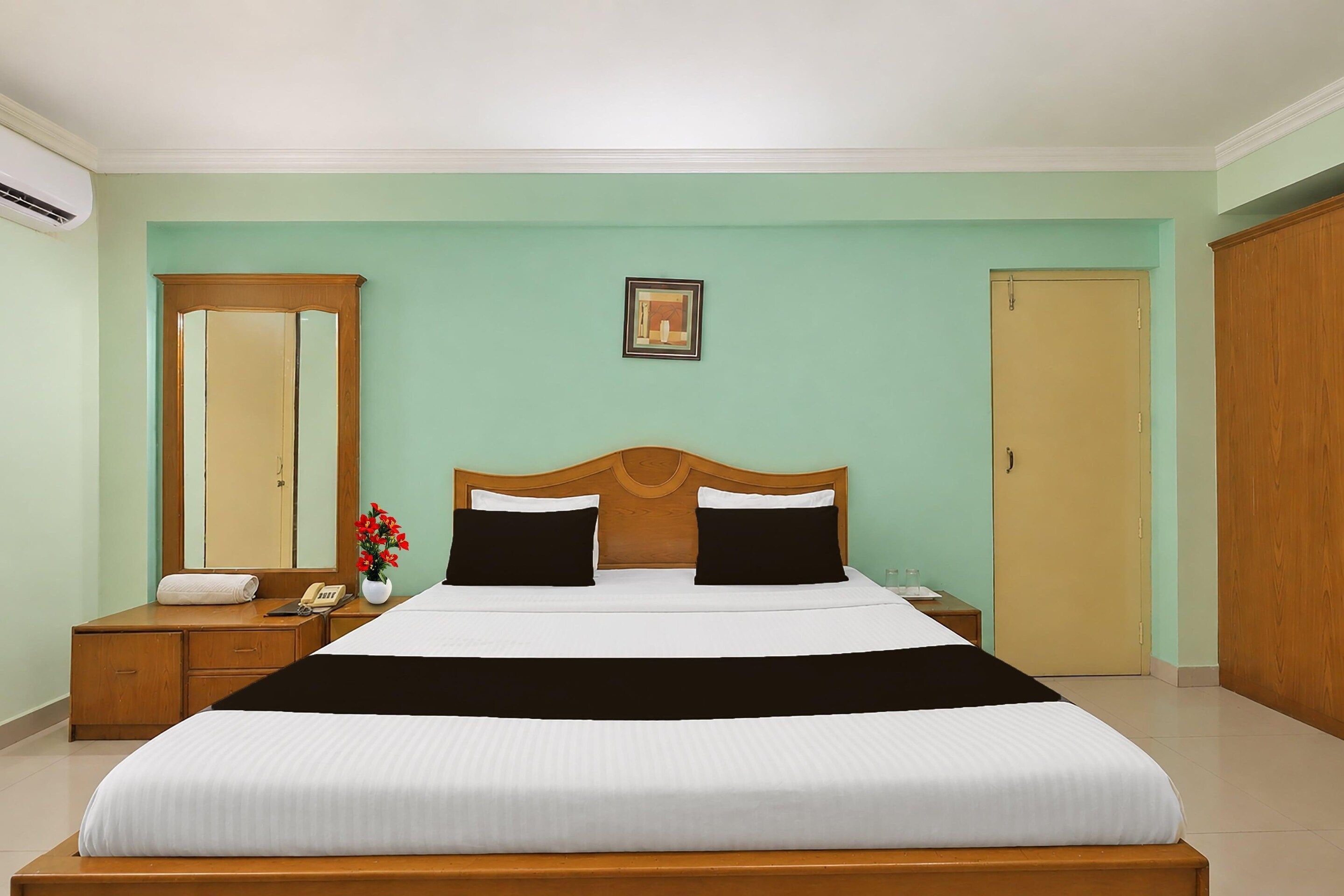 A hotel room with a bed, a bedside table, a mirror, a picture on the wall, and a door.