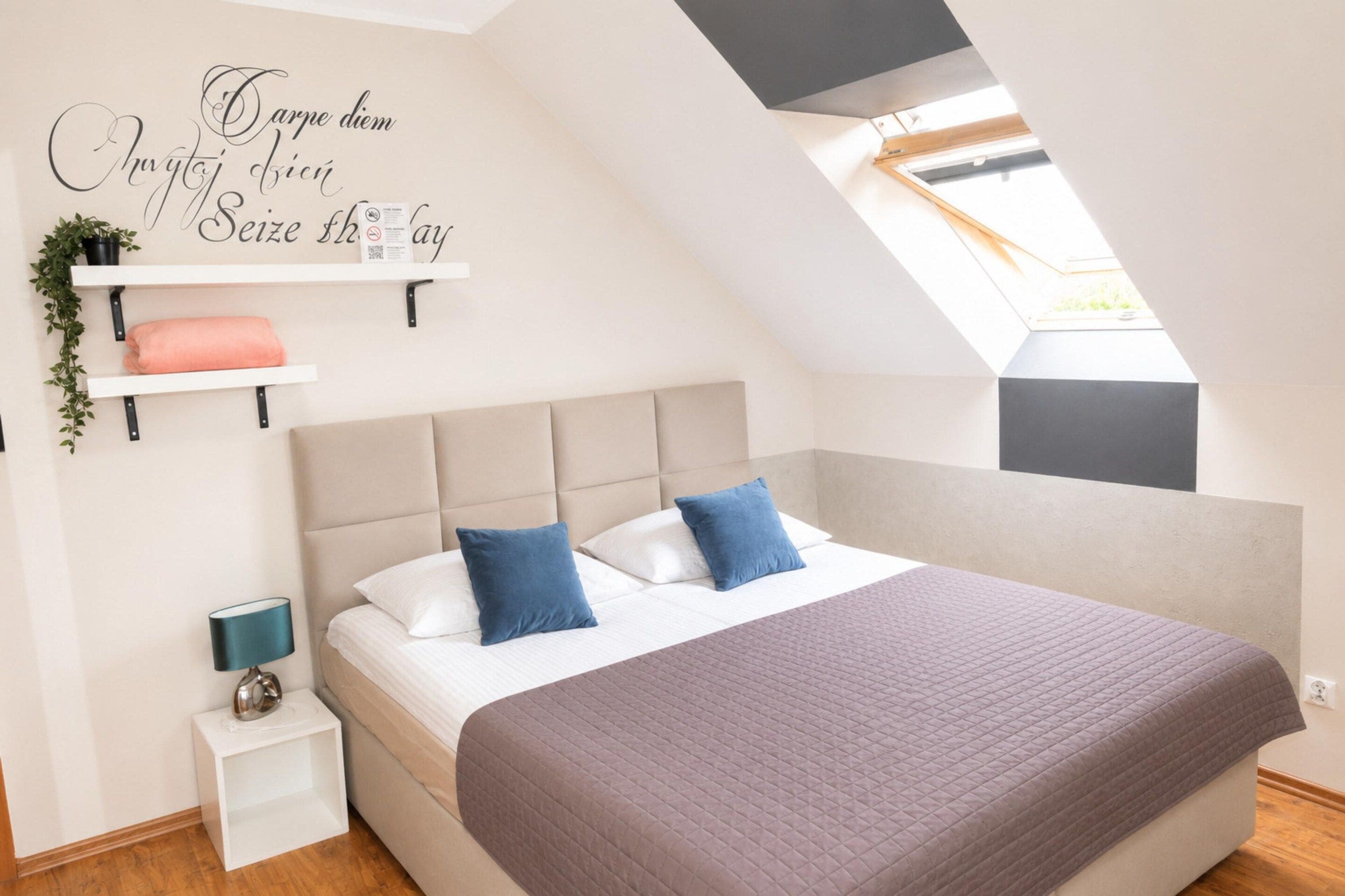 A bedroom with a sloped ceiling, a bed with two pillows, a bedside table with a lamp, and a wall-mounted shelf with a plant.