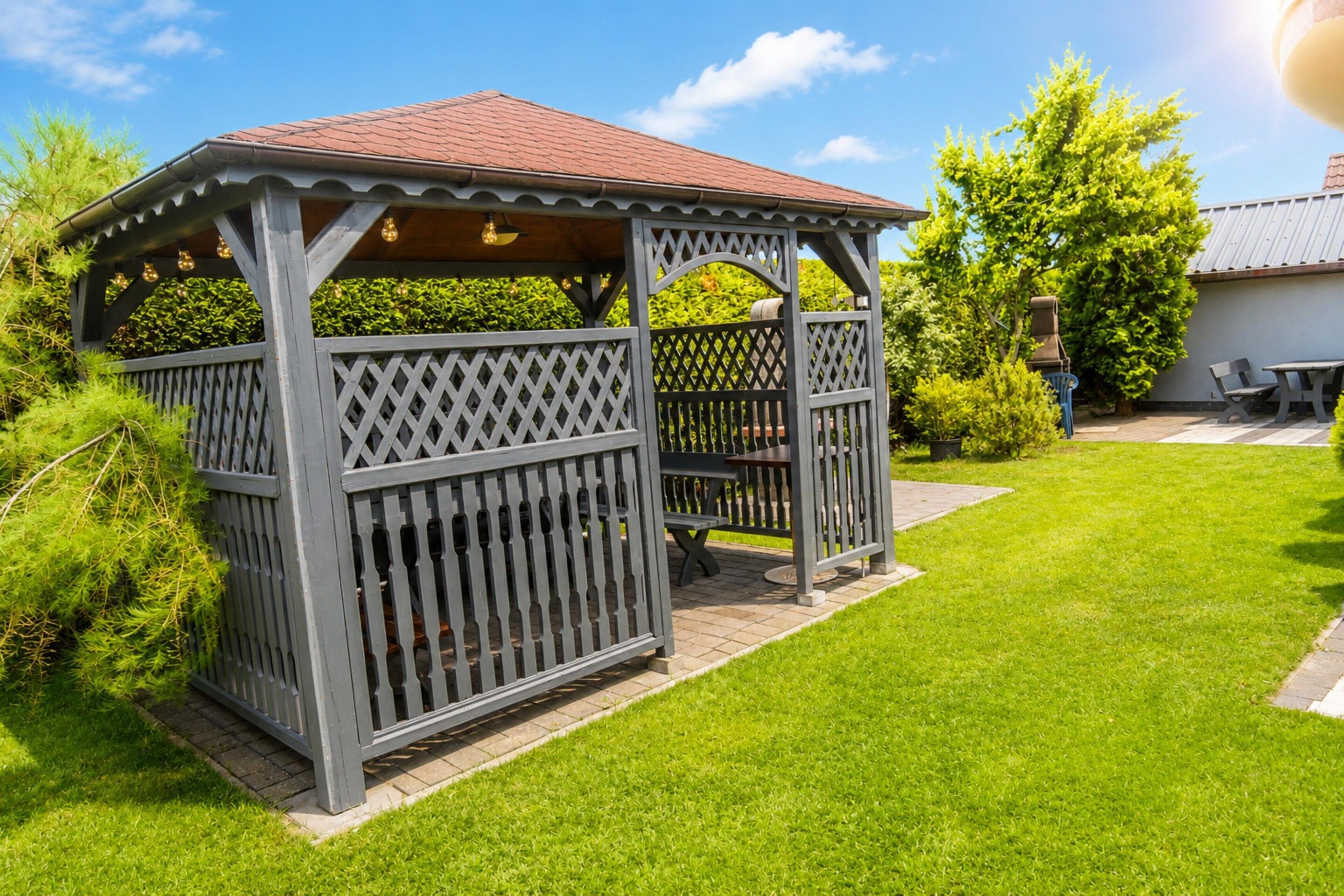 A wooden gazebo with a lattice design, a bench, and a table, surrounded by a well-maintained lawn and trees.