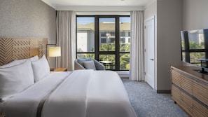 Ein Hotelzimmer mit einem großen Bett, einem Schreibtisch, einem Sessel, einem Fernseher und einem Balkon mit Blick auf Gebäude und Palmen.