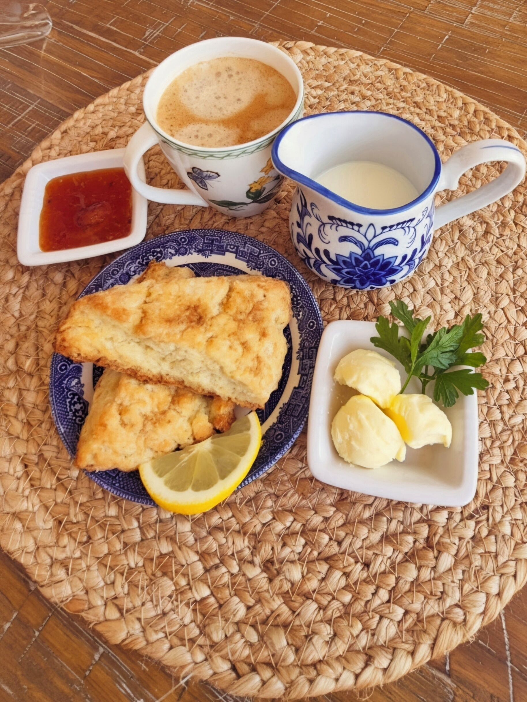 A table setting with a woven placemat, a plate with two pieces of fried food, a lemon wedge, a small bowl of butter, a cup of coffee, and a small bowl of sauce.