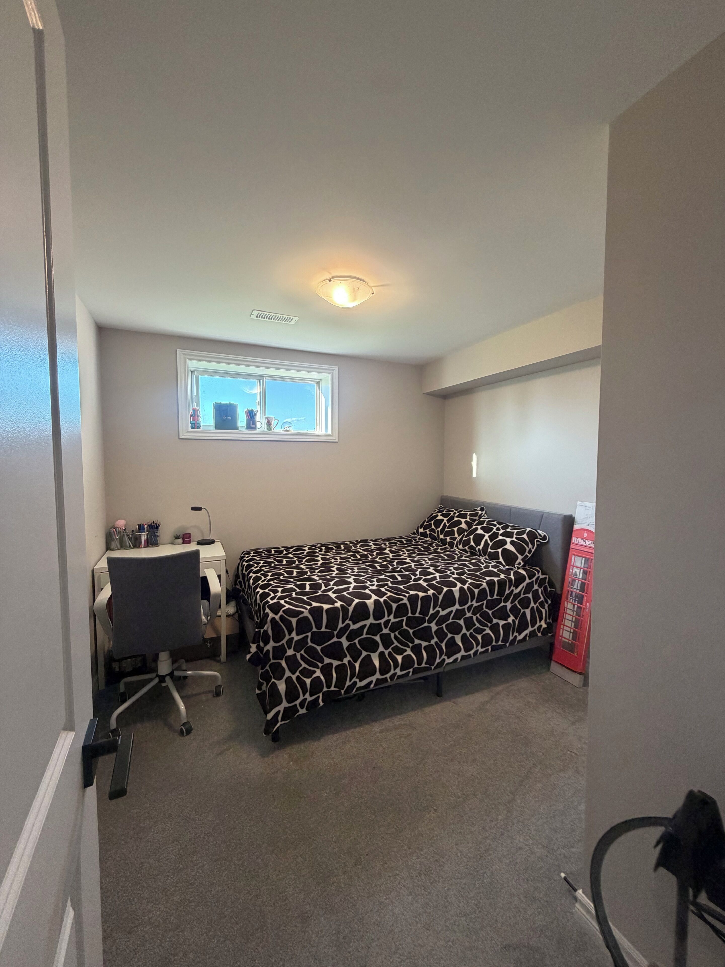 A bedroom with a bed, a desk, a chair, a window, and a red telephone booth.