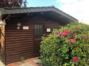 A wooden building with a 'Sauna' sign, a window with a decorative grille, and a bush with pink flowers. - A cozy retreat for a getaway by the sea; dogs welcome—with a pool and sauna (Butjadingen)
