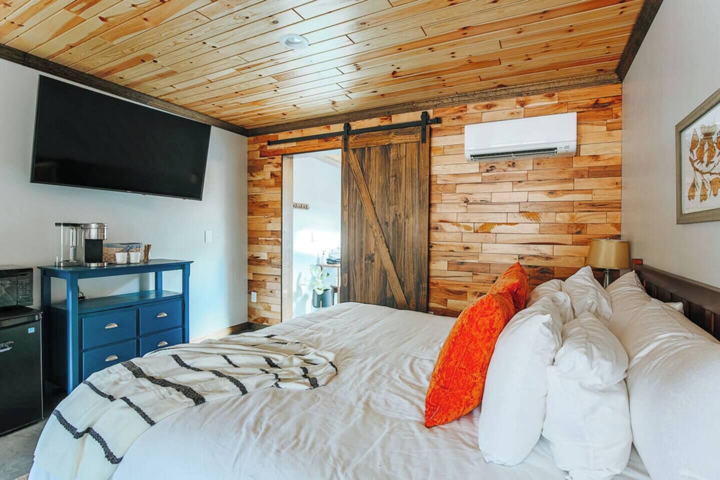 Un dormitorio con techo de madera, una cama con almohadas blancas y anaranjadas, un aparador azul y un televisor de pantalla plana montado en la pared.