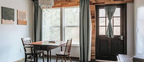 A dining area with a wooden table and chairs, a chandelier, and a view of the outdoors through a window.