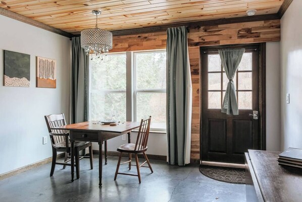 A dining area with a wooden table and chairs, a chandelier, and a view of the outdoors through a window.