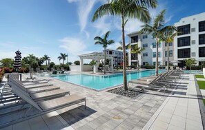 Uma piscina com espreguiçadeiras, palmeiras e um edifício moderno ao fundo.