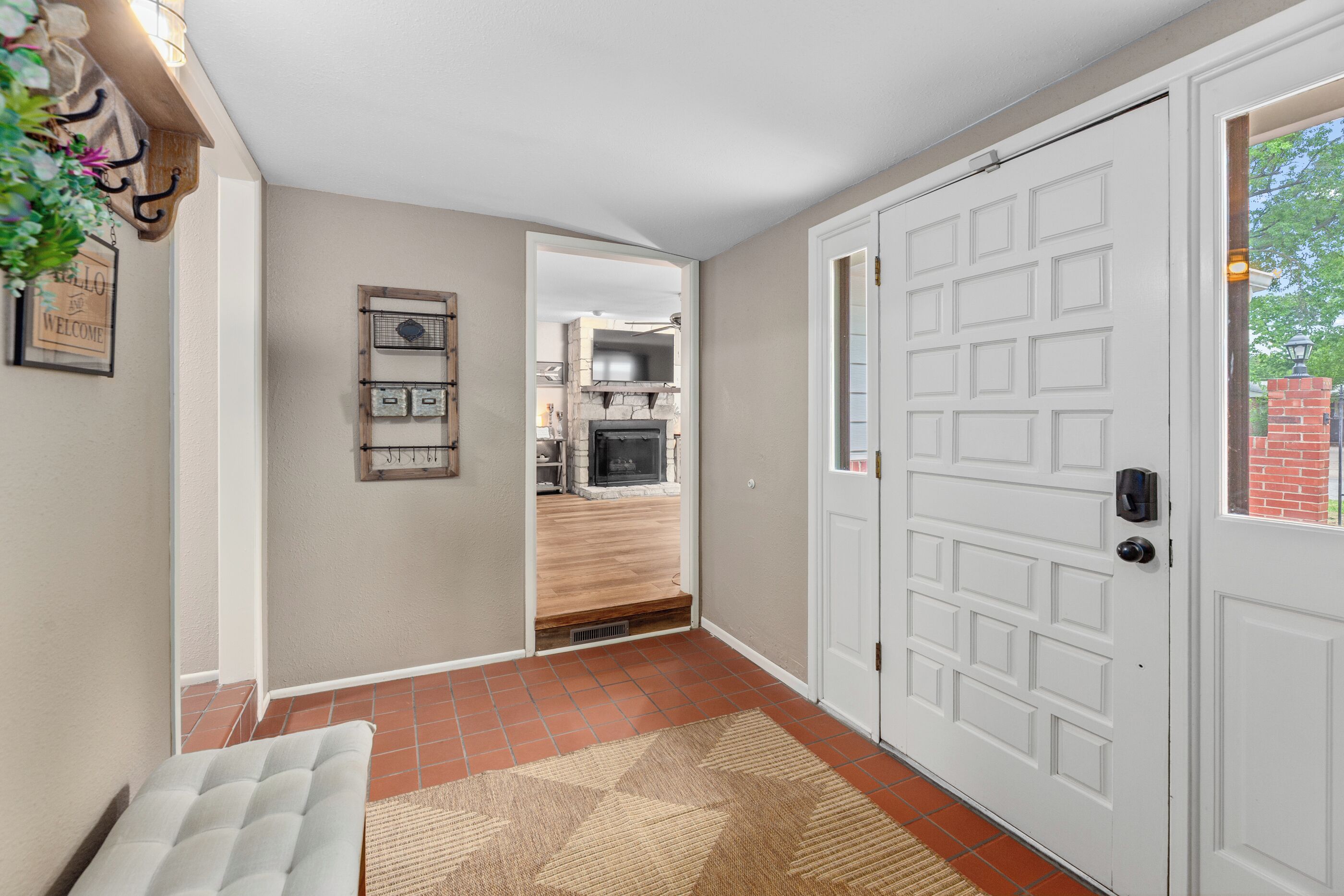 A hallway with a white door, a bench, a welcome sign, and a view into a kitchen area.