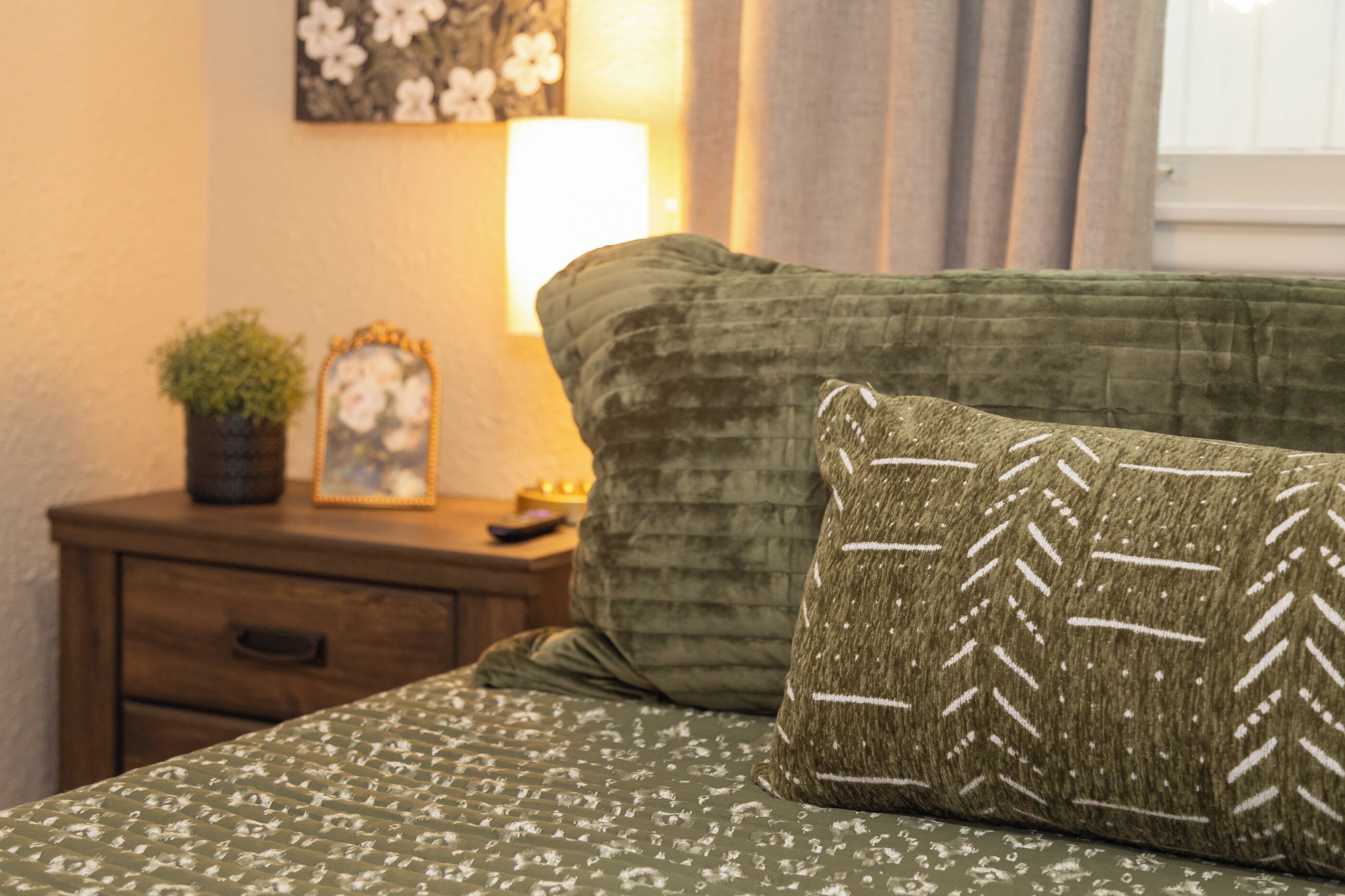 Ein Bett mit gemustertem Überwurf und einem Kopfkissen mit geometrischem Muster, ein Nachttisch mit Lampe, ein gerahmtes Bild und ein Blumentopf.