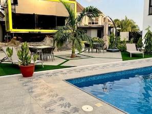 Una piscina con solárium de baldosas, una palmera y una zona de estar al aire libre techada.