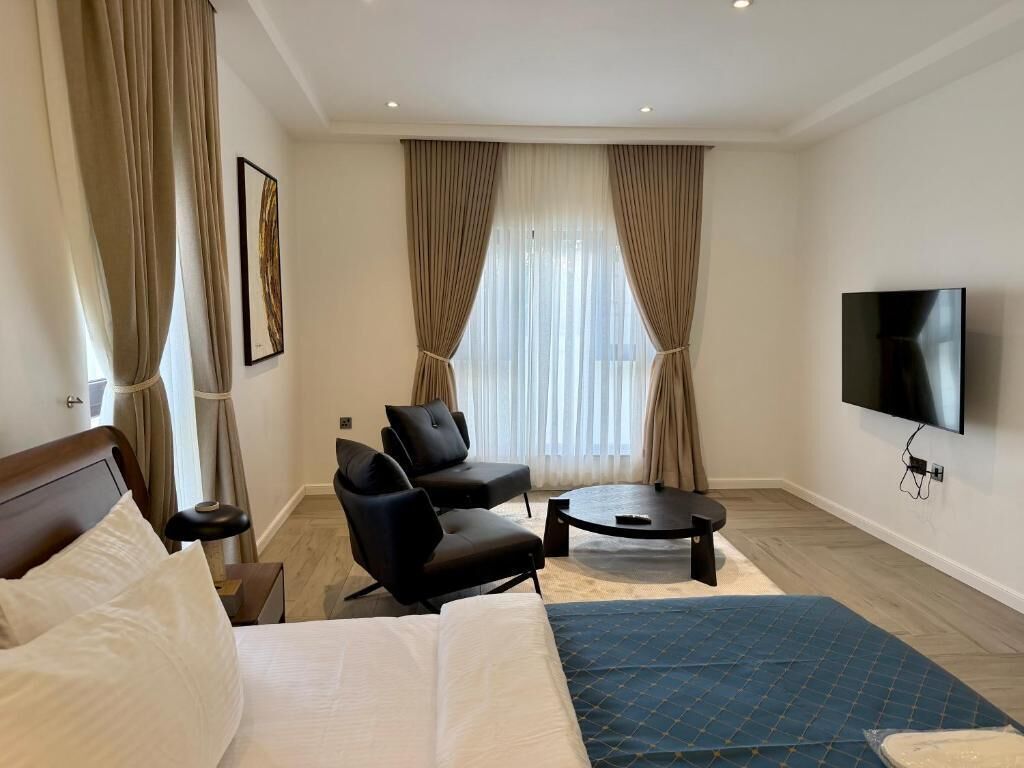 A hotel room with a bed, two armchairs, a coffee table, a TV, and a large window with curtains.