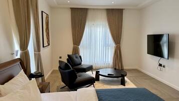 A hotel room with a bed, two armchairs, a coffee table, a TV, and a large window with curtains.