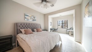 Ein Schlafzimmer mit einem karabinierten Kopfteil, einem Bett mit weißer und beiger Bettdecke, einem Nachttisch und einem Deckenventilator.