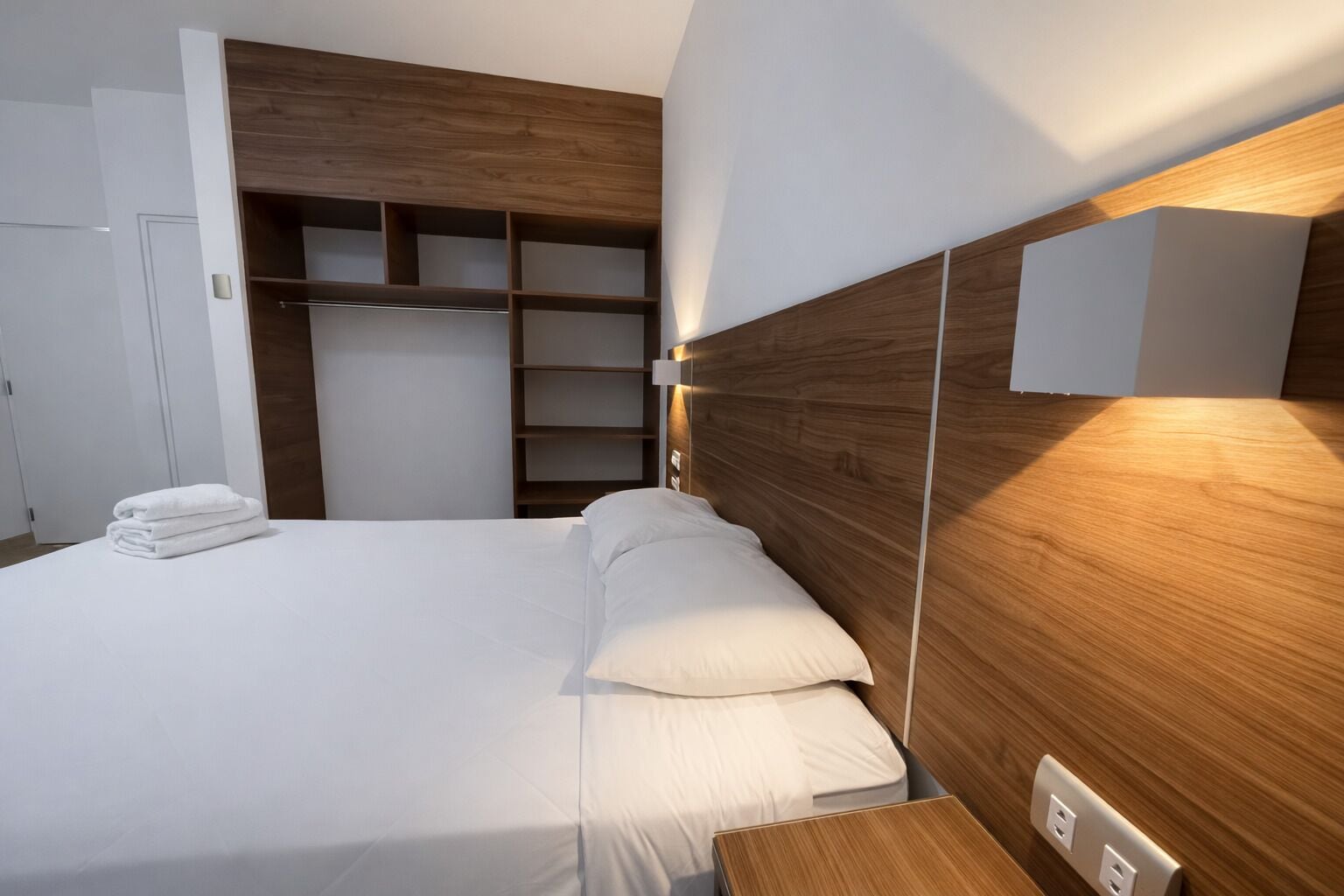 Une chambre d’hôtel moderne avec un lit bien fait, une tête de lit en bois, une armoire intégrée et une table de chevet avec une lampe.