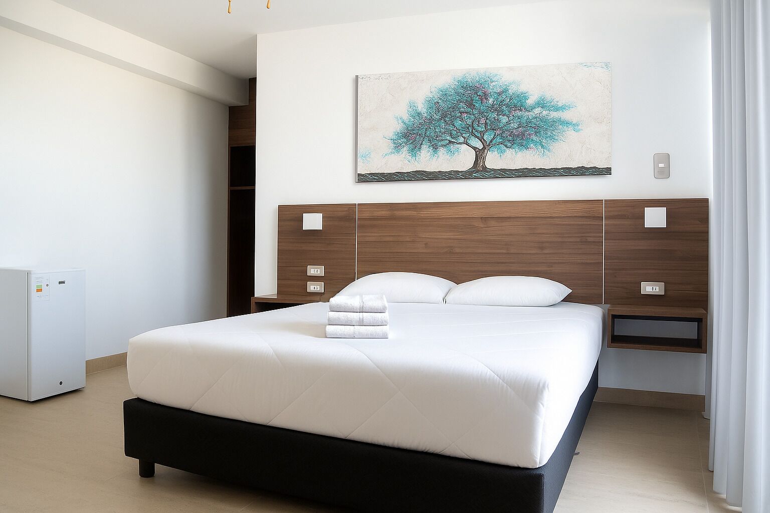 A modern hotel room with a large bed, a wooden headboard, a nightstand, a wall-mounted painting, and a built-in wardrobe.