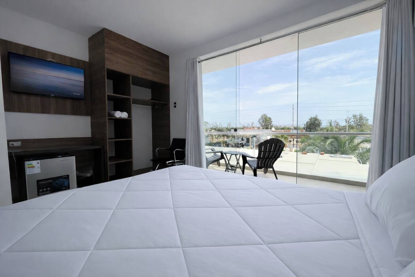 Habitación de hotel con cama, televisión de pantalla plana, minibar, zona de estar con mesa y sillas, y un ventanal con vistas al exterior.