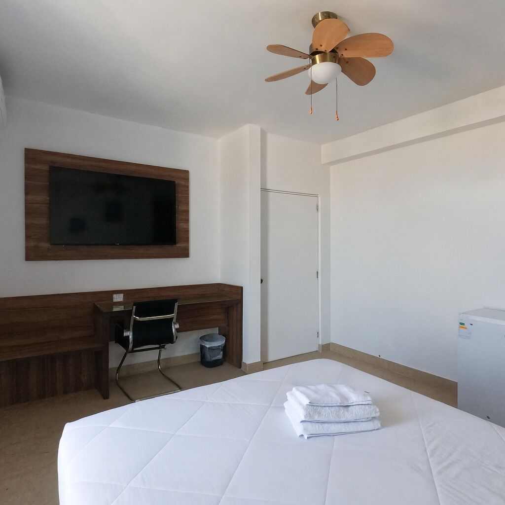 A hotel room with a bed, a desk, a television mounted on the wall, and a ceiling fan.