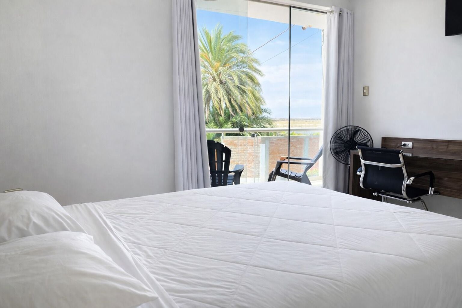 Habitación de hotel con una cama grande, una silla, un ventilador y vistas a palmeras a través de una puerta corredera.