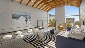 Een modern interieur met een hoge plafond, een groot schilderij van een vuurtoren, een zithoek met kussens, en een geruit vloerkleed.