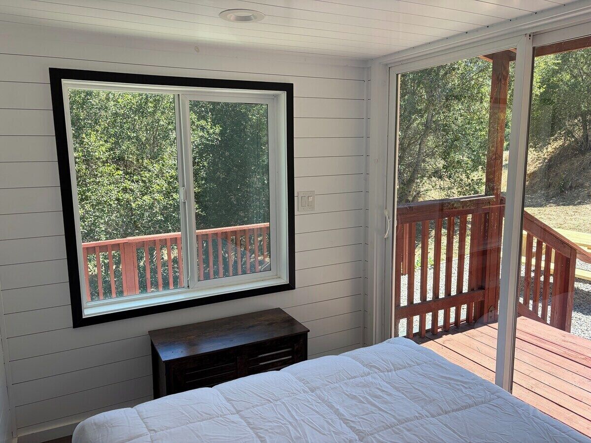A bedroom with a large window, a bed, a nightstand, and a view of a deck and trees.